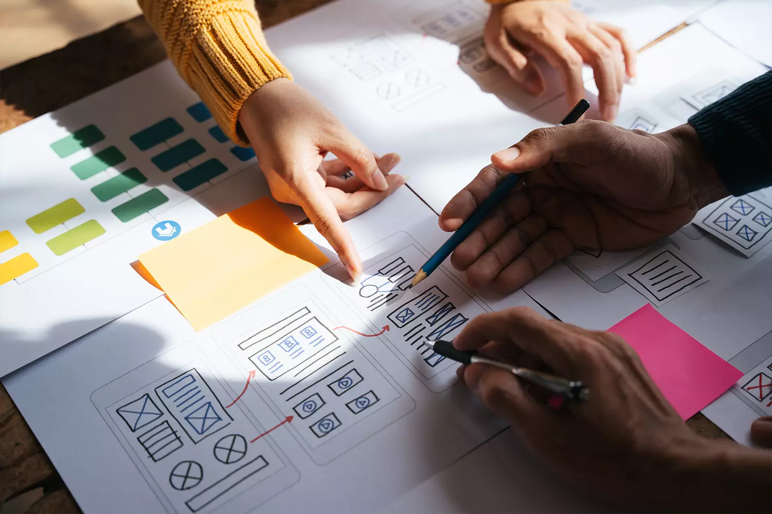 Close-up of hands working on a project with diagrams and sticky notes. Several fingers point at different parts of the papers on the table, indicating collaboration and User Experience planning.