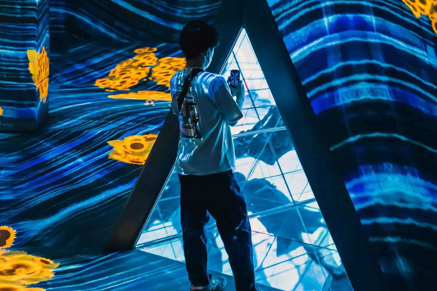 A person stands inside a triangular structure taking a photo, surrounded by vibrant, abstract digital art featuring swirling blue patterns and bright yellow flowers, reminiscent of innovative website design aesthetics.