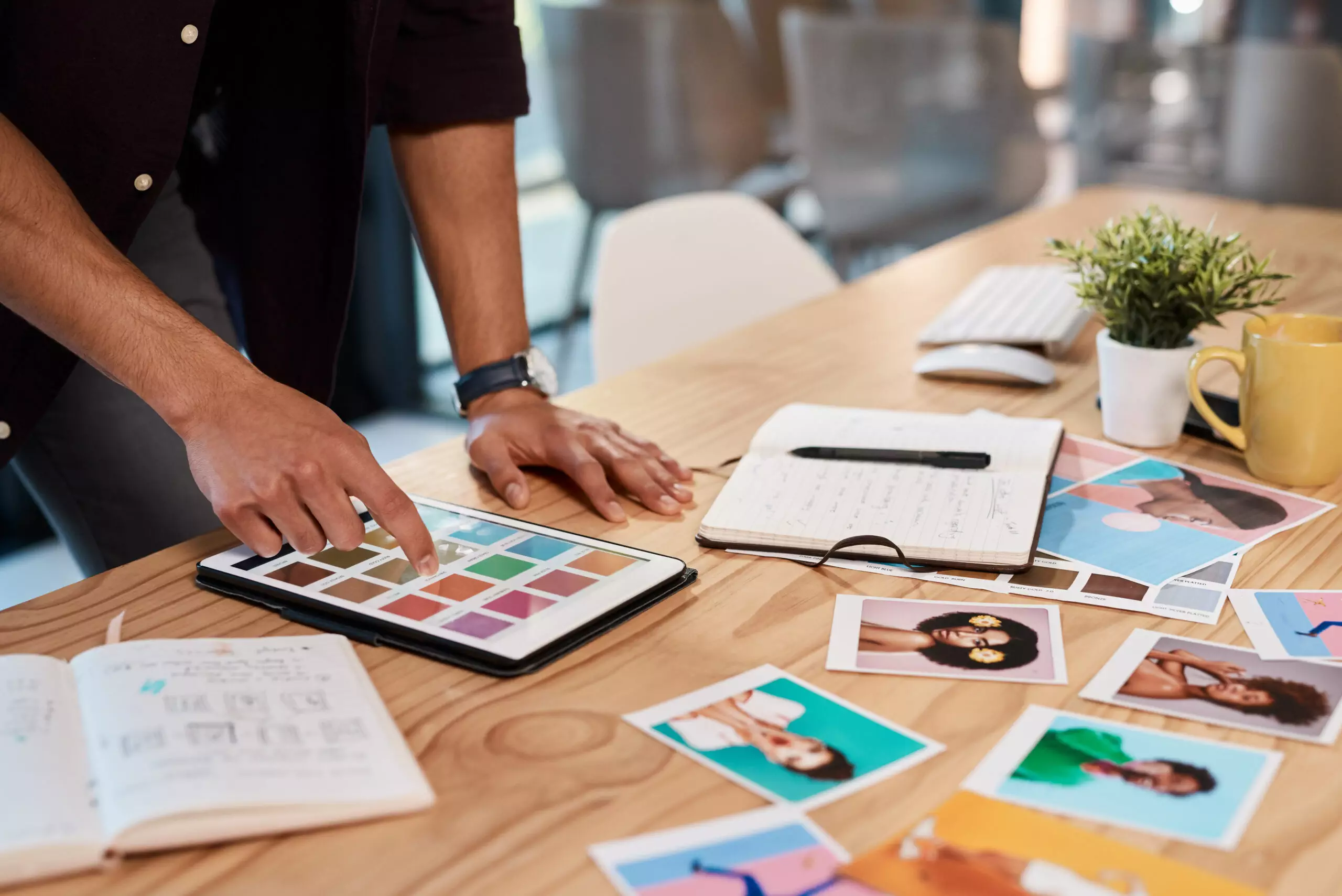 A person engaged in art direction uses a tablet to select colors at a desk, surrounded by notebooks, printed photos, a plant, and a coffee cup.