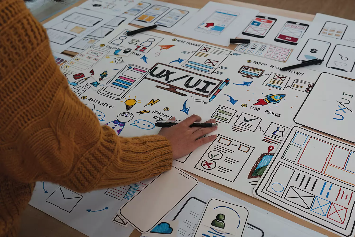 A person in a yellow sweater is working diligently on various UX/UI design sketches and wireframes, integral aspects of digital product development, spread across the table.